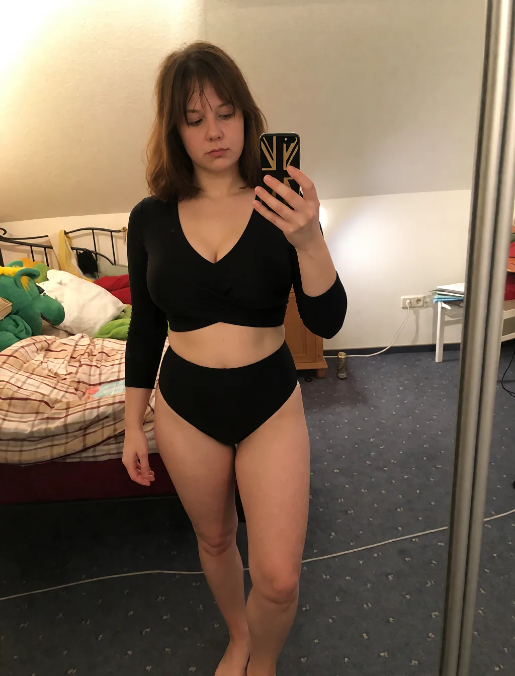 Edit the supplied photograph. Change only what is asked for below. Return one clean, natural, high-resolution photograph with realistic skin texture and no distortion. Make her chest noticeably larger and fuller, in a natural shape that suits her build, and let whatever she is wearing fall over it correctly. Replace her outfit with a fitted two-piece swimsuit that suits her. Keep her face, her identity, her hair and her skin tone exactly as they are, and keep the room and the light she is in. Keep her pose and the framing of the shot.