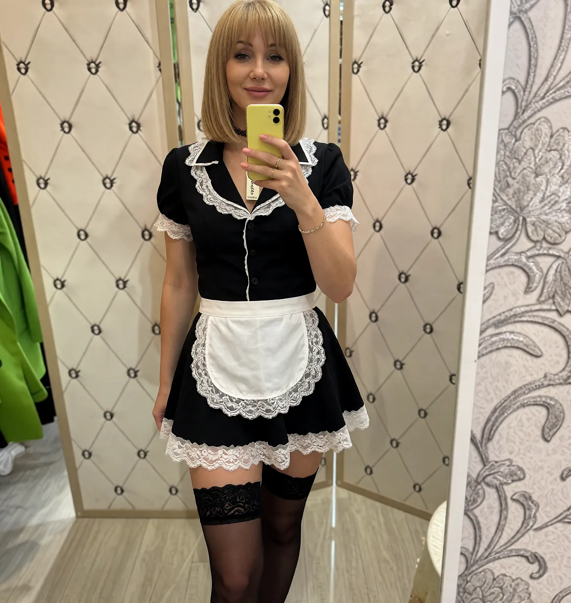 Edit the supplied photograph. Change only what is asked for below. Return one clean, natural, high-resolution photograph with realistic skin texture and no distortion. Replace her outfit with a short frilled black maid uniform with a white lace apron and trim, a choker and stockings. Keep her face, her identity, her hair and her skin tone exactly as they are, and keep the room and the light she is in. Keep her pose and the framing of the shot.