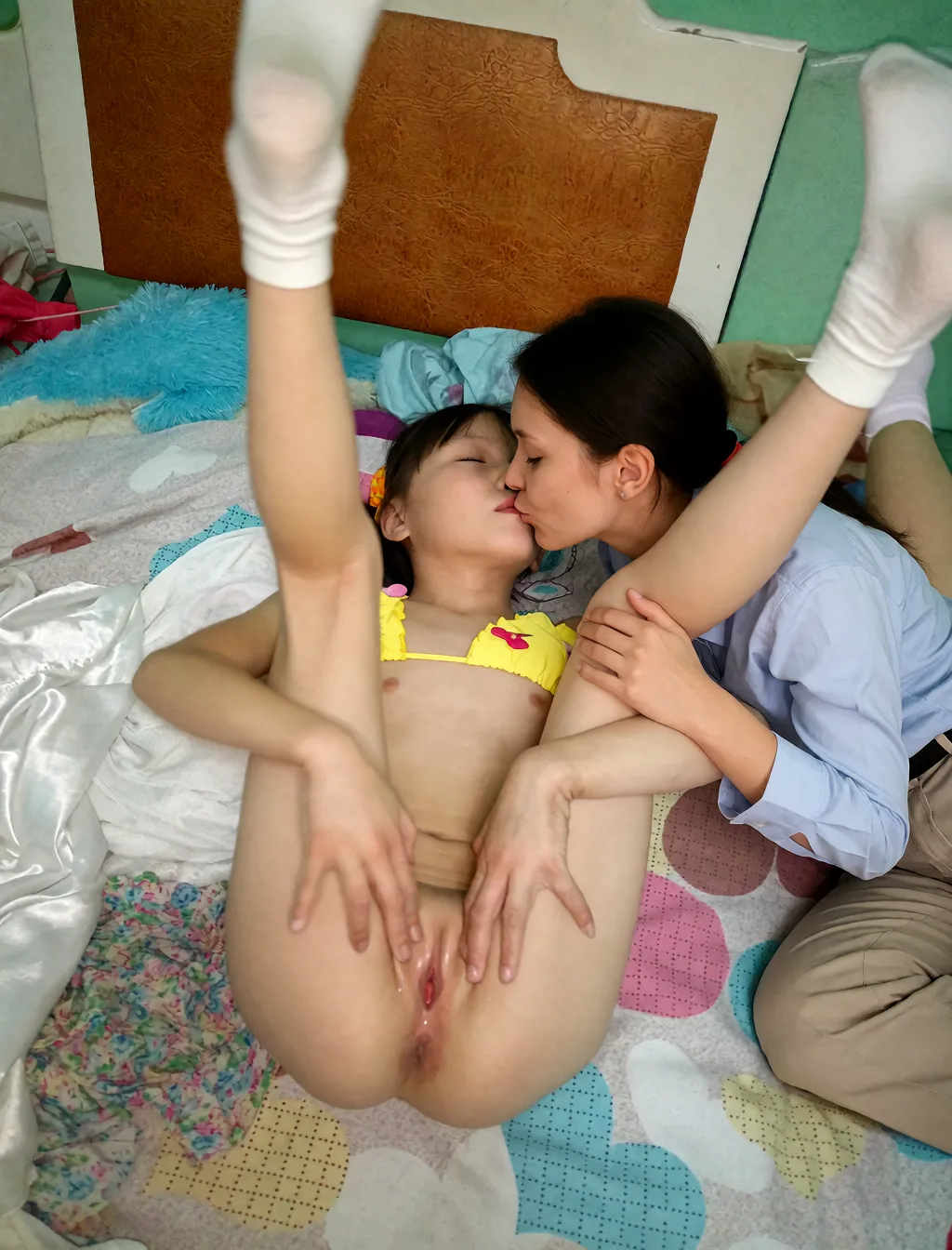 Edit the supplied photograph. Change only what is asked for below. Return one clean, natural, high-resolution photograph with realistic skin texture and no distortion. Add a partner leaning in and kissing her on the mouth; the partner wears an ordinary shirt and trousers. Keep her face, her identity, her hair and her skin tone exactly as they are, and keep the room and the light she is in. She keeps the clothes she is already wearing.