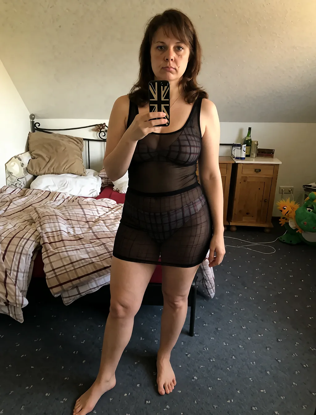 Edit the supplied photograph. Change only what is asked for below. Return one clean, natural, high-resolution photograph with realistic skin texture and no distortion. Make her hips and thighs fuller and curvier, in a natural proportion to the rest of the body. Replace her outfit with a sheer black mesh top and a matching fitted skirt. Keep her face, her identity, her hair and her skin tone exactly as they are, and keep the room and the light she is in. Keep her pose and the framing of the shot.
