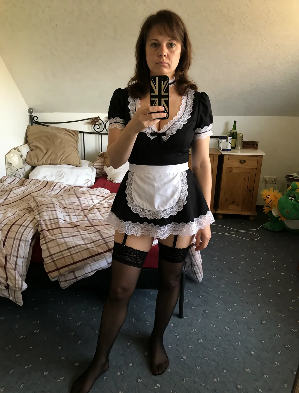 Edit the supplied photograph. Change only what is asked for below. Return one clean, natural, high-resolution photograph with realistic skin texture and no distortion. Replace her outfit with a short frilled black maid uniform with a white lace apron and trim, a choker and stockings. Keep her face, her identity, her hair and her skin tone exactly as they are, and keep the room and the light she is in. Keep her pose and the framing of the shot.