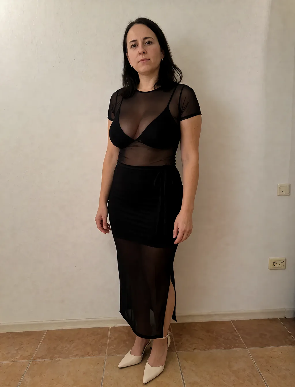 Edit the supplied photograph. Change only what is asked for below. Return one clean, natural, high-resolution photograph with realistic skin texture and no distortion. Make her chest noticeably larger and fuller, in a natural shape that suits her build, and let whatever she is wearing fall over it correctly. Replace her outfit with a sheer black mesh top and a matching fitted skirt. Keep her face, her identity, her hair and her skin tone exactly as they are, and keep the room and the light she is in. Keep her pose and the framing of the shot.