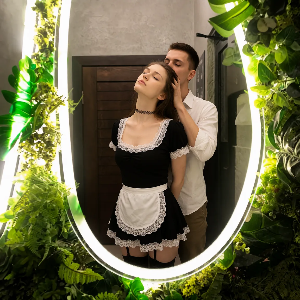 Edit the supplied photograph. Change only what is asked for below. Return one clean, natural, high-resolution photograph with realistic skin texture and no distortion. Make her chest noticeably smaller, in a natural shape that suits her build, and let whatever she is wearing fall over it correctly. Replace her outfit with a short frilled black maid uniform with a white lace apron and trim, a choker and stockings. Add a partner standing behind her with one hand gathered in her hair, tilting her head back so her throat is bared while she closes her eyes; the partner wears an ordinary shirt and trousers. Keep her face, her identity, her hair and her skin tone exactly as they are, and keep the room and the light she is in.