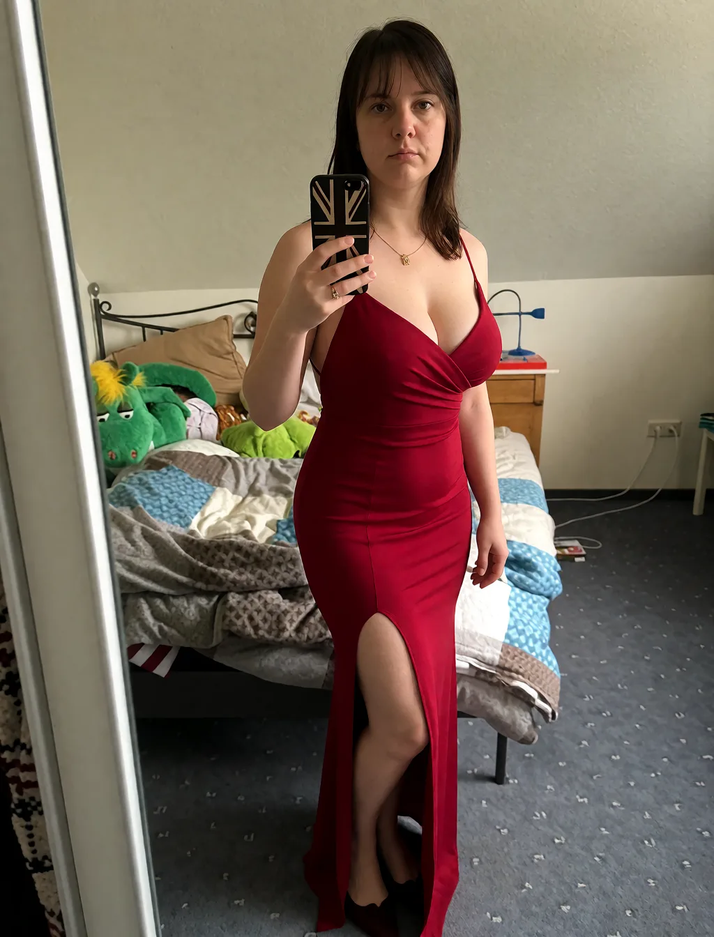 Edit the supplied photograph. Change only what is asked for below. Return one clean, natural, high-resolution photograph with realistic skin texture and no distortion. Make her chest noticeably larger and fuller, in a natural shape that suits her build, and let whatever she is wearing fall over it correctly. Replace her outfit with a fitted red evening dress with a high slit and a low back. Keep her face, her identity, her hair and her skin tone exactly as they are, and keep the room and the light she is in. Keep her pose and the framing of the shot.