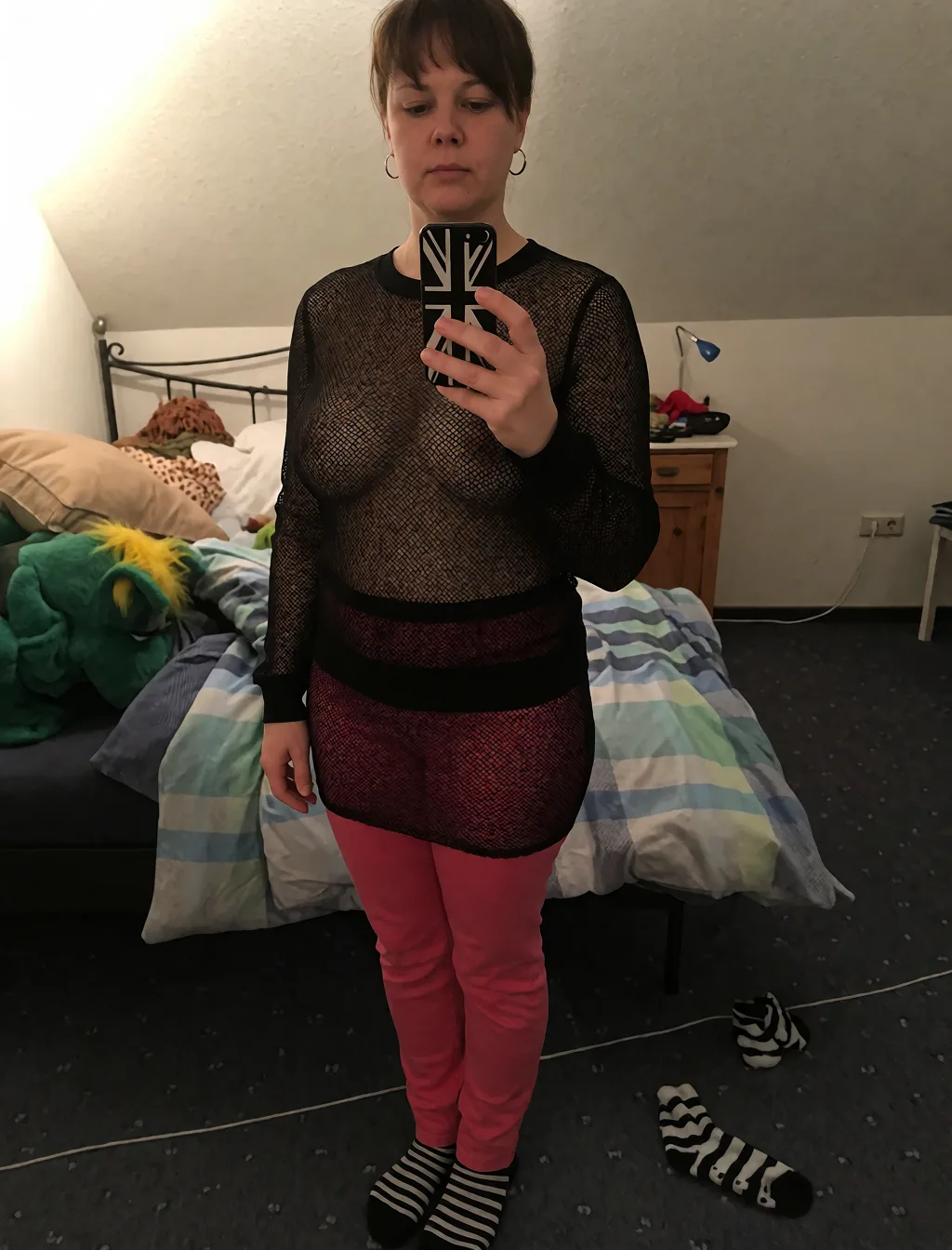 Edit the supplied photograph. Change only what is asked for below. Return one clean, natural, high-resolution photograph with realistic skin texture and no distortion. Make her chest noticeably larger and fuller, in a natural shape that suits her build, and let whatever she is wearing fall over it correctly. Replace her outfit with a sheer black mesh top and a matching fitted skirt. Keep her face, her identity, her hair and her skin tone exactly as they are, and keep the room and the light she is in. Keep her pose and the framing of the shot.