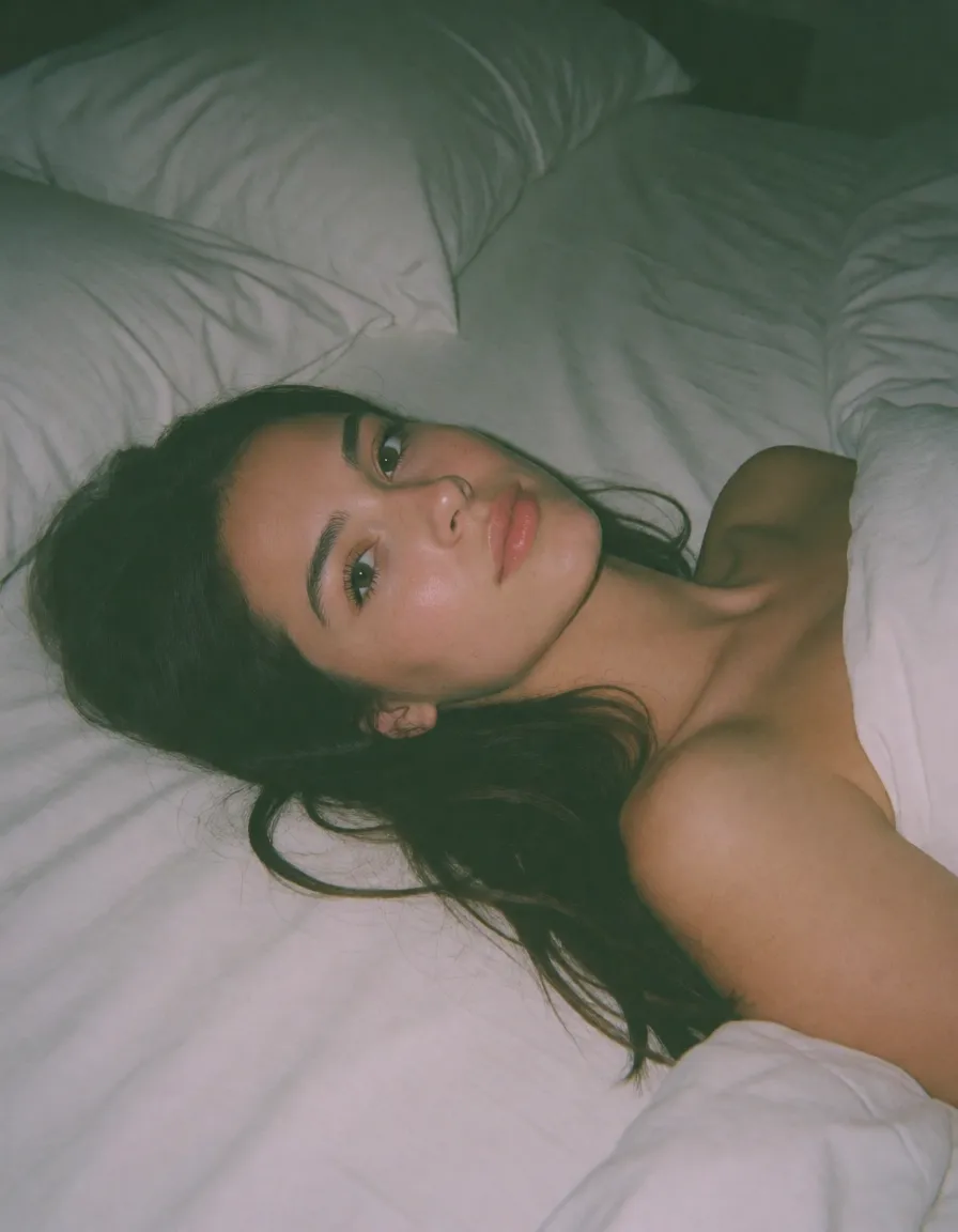 an amateur snapshot, a nude woman with long dark hair lying back on white sheets, legs apart, looking at the viewer, dim flat indoor light, washed out muted colour, faint green cast, visible noise, plain unpolished snapshot, natural unretouched skin, strikingly beautiful, radiant complexion, early twenties