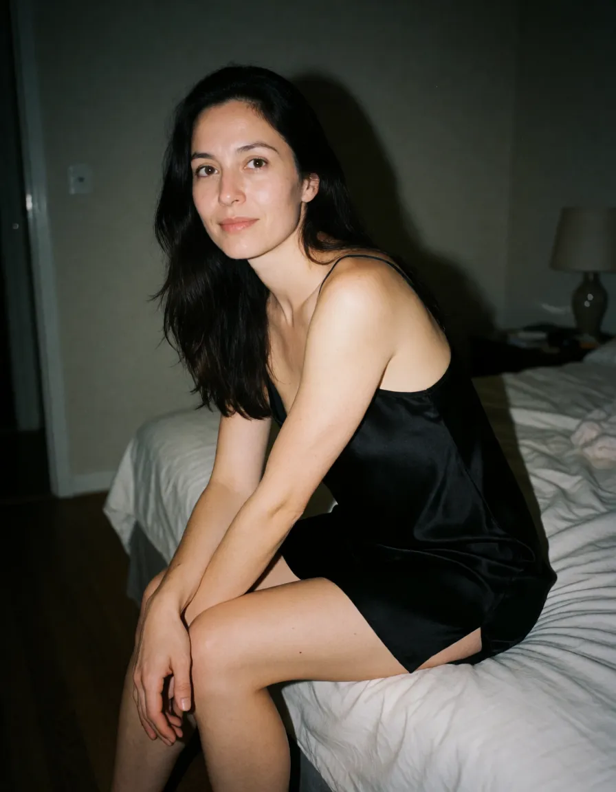 an amateur homemade snapshot, real ordinary photo, a woman with long dark hair in a black silk slip, sitting on the edge of a bed, looking at the viewer, shot on a phone with the flash on, harsh direct flash falling flat on the skin, bright hot subject with the background dropping into darkness behind her, completely unretouched real skin with visible pores, freckles, moles, stray hairs and small blemishes, no makeup, no filter, plain messy everyday room, slightly soft handheld focus, digital sensor noise, crooked casual framing